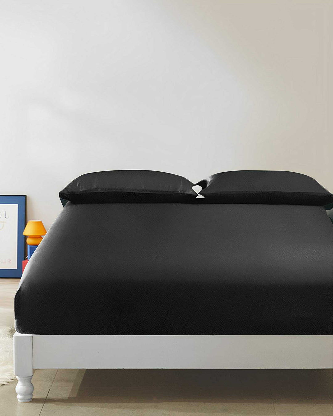 A neatly made bed with luxurious SusanSilk 19 Momme Seamless Silk Fitted Sheet in black and two matching pillows, positioned in a minimalist bedroom with white walls and simple decor items visible in the background.