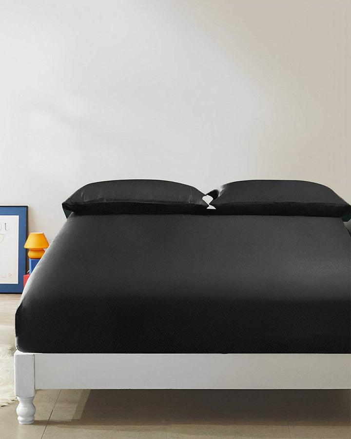 A neatly made bed with luxurious SusanSilk 19 Momme Seamless Silk Fitted Sheet in black and two matching pillows, positioned in a minimalist bedroom with white walls and simple decor items visible in the background.