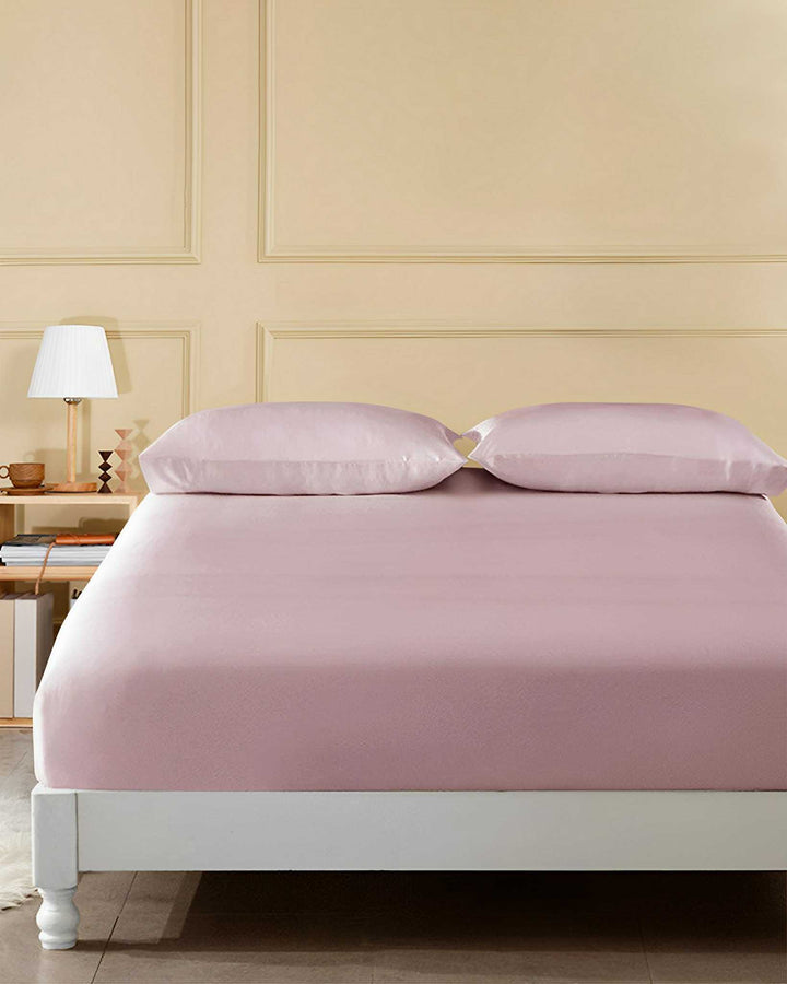 A neatly made bed adorned with a 19 Momme Seamless Silk Fitted Sheet from SusanSilk in light pink and two pillows is positioned against a cream-colored wall with panel detailing. On the left side, there is a nightstand holding a lamp and several books.