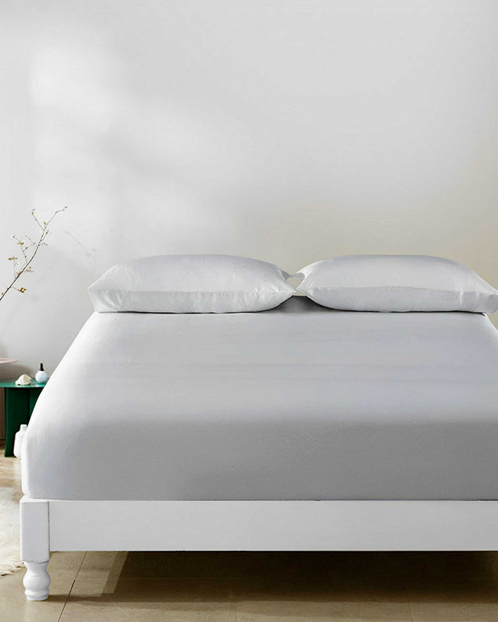 A neatly made bed featuring SusanSilk's 19 Momme Seamless Silk Fitted Sheet and a white duvet with two pillows, set against a plain white wall. A small green nightstand with a vase and a bottle is visible in the background.
