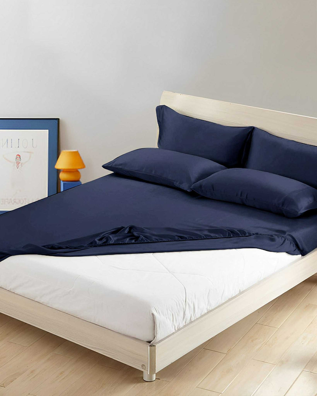 A neatly made bed featuring a 19 Momme Seamless Silk Fitted Sheet by SusanSilk in dark blue, accompanied by matching pillows, is situated in a minimalist room with light wood flooring. A poster and an orange lamp can be seen in the background.