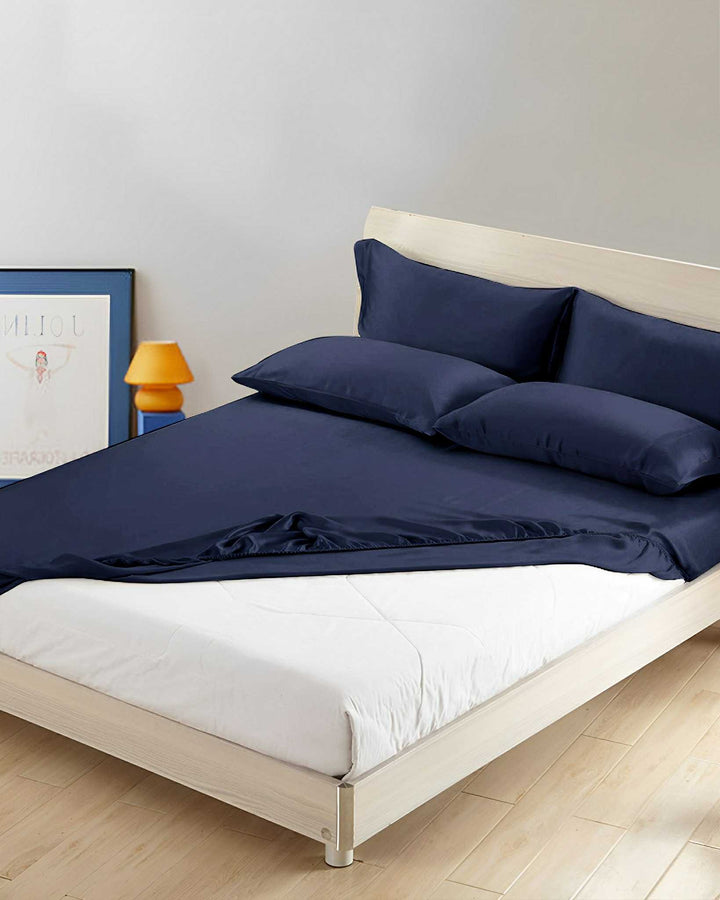 A neatly made bed featuring a 19 Momme Seamless Silk Fitted Sheet by SusanSilk in dark blue, accompanied by matching pillows, is situated in a minimalist room with light wood flooring. A poster and an orange lamp can be seen in the background.