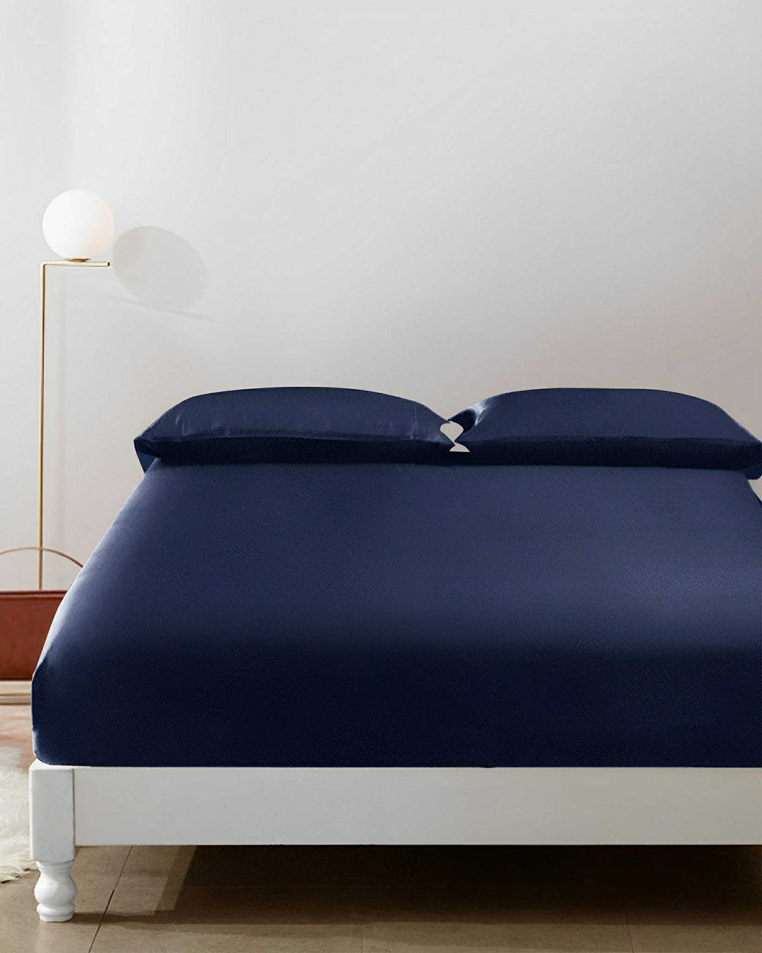 A neatly made bed with dark blue 19 Momme Seamless Silk Fitted Sheets by SusanSilk and matching pillows stands against a plain white wall. A round white lamp on a tall stand is positioned on the left side, with a small red table below it.