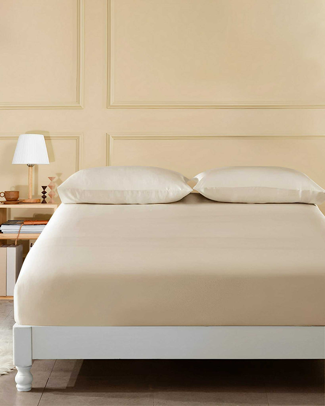 A neatly made bed with SusanSilk's 19 Momme Seamless Silk Fitted Sheet in a soft beige, paired with matching pillows, sits beside a nightstand adorned with a lamp and books against a light beige paneled wall.