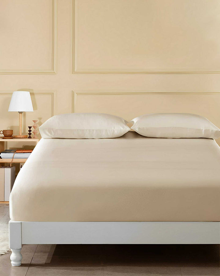 A neatly made bed with SusanSilk's 19 Momme Seamless Silk Fitted Sheet in a soft beige, paired with matching pillows, sits beside a nightstand adorned with a lamp and books against a light beige paneled wall.