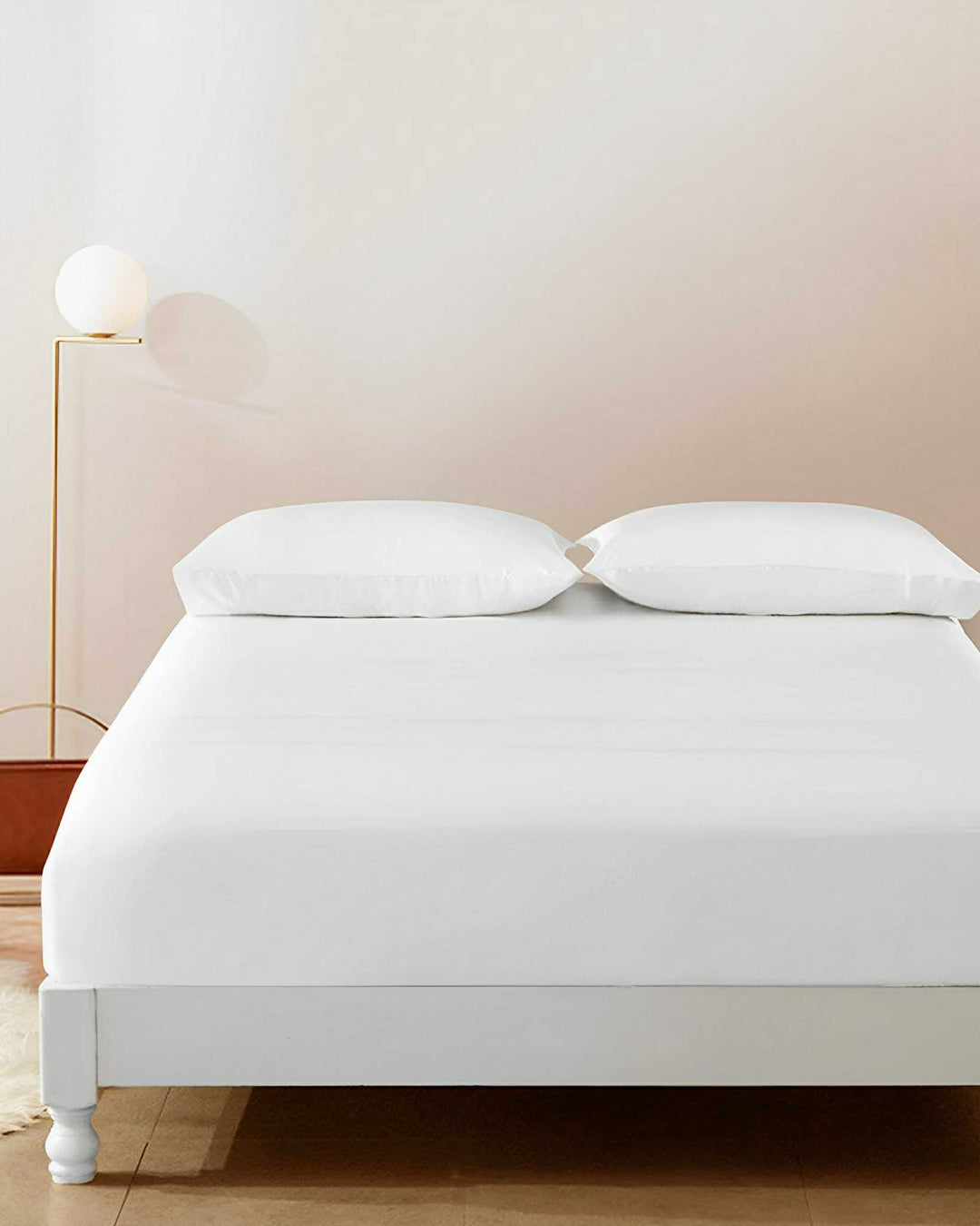 A neatly made bed with a 19 Momme Seamless Silk Fitted Sheet by SusanSilk, two white pillows, and a sleek white headboard. A modern floor lamp with a globe-shaped light stands beside the bed on a wooden floor.