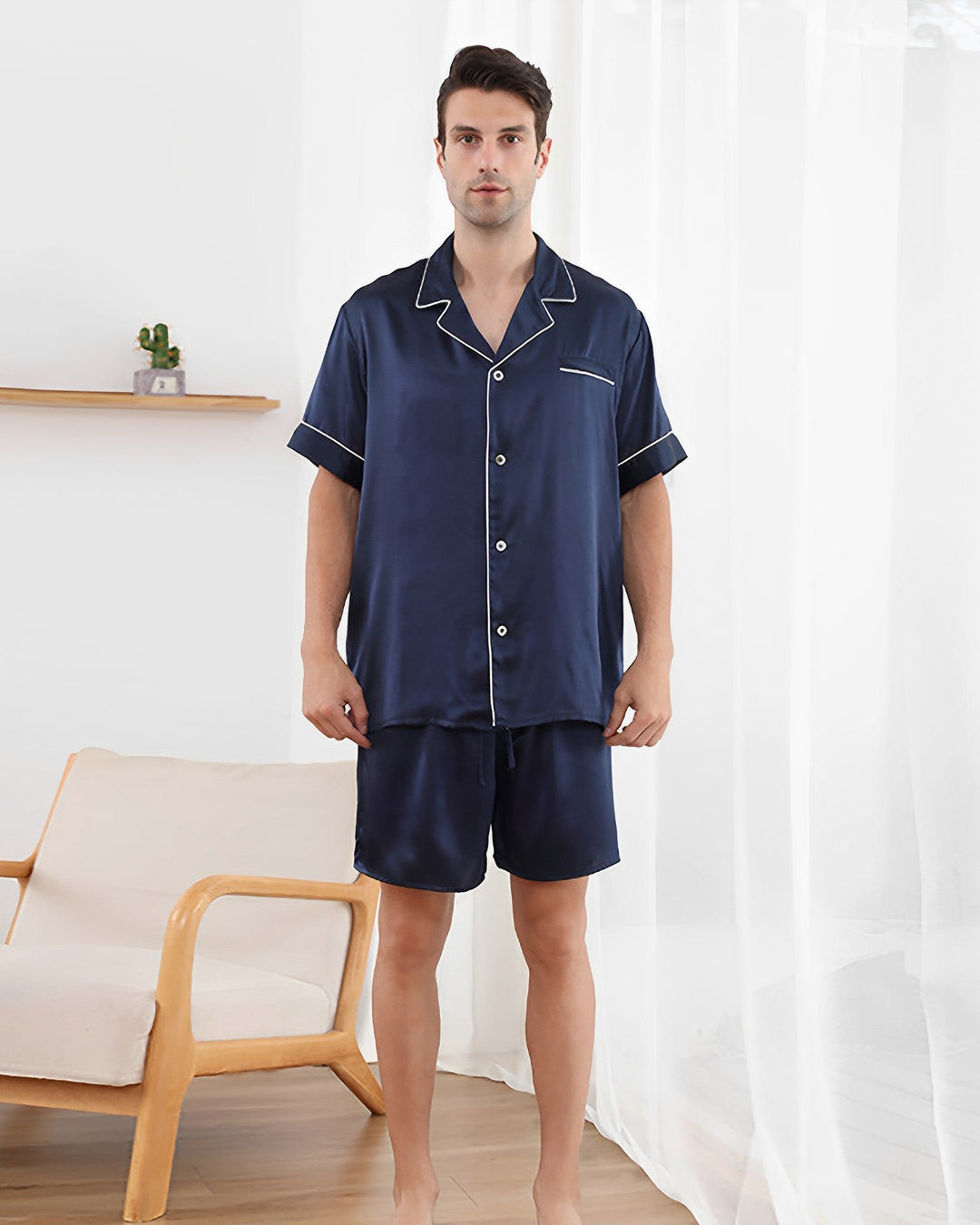 A man stands indoors wearing a 19 Momme Short Silk Pajamas Set by SusanSilk in navy blue. To his left is a wooden chair, and the back wall features a small shelf adorned with a potted plant. The windows are covered with sheer curtains.