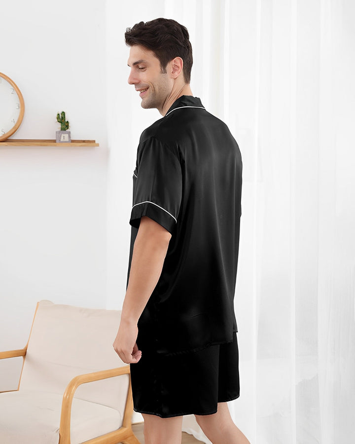 A man with short dark hair stands in a bright room, wearing the SusanSilk 19 Momme Short Silk Pajamas Set in sleek black, and facing partially away from the camera. A clock and small plant are visible on a wall shelf in the background.