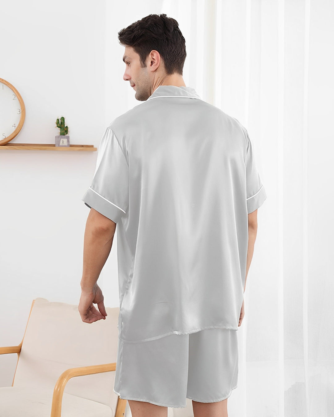 A man wearing the light gray 19 Momme Short Silk Pajamas Set by SusanSilk is seen from the back, standing in a room with light-colored decor, including a small wall clock and a potted cactus on a shelf. The luxurious sheen of the fabric confirms it is made of silk.