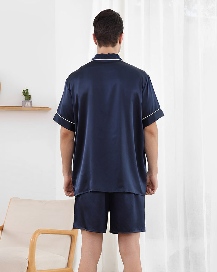 A person stands with their back facing the camera, wearing the 19 Momme Short Silk Pajamas Set in dark blue from SusanSilk. They are in a room with a beige chair and a small shelf hosting a cactus.