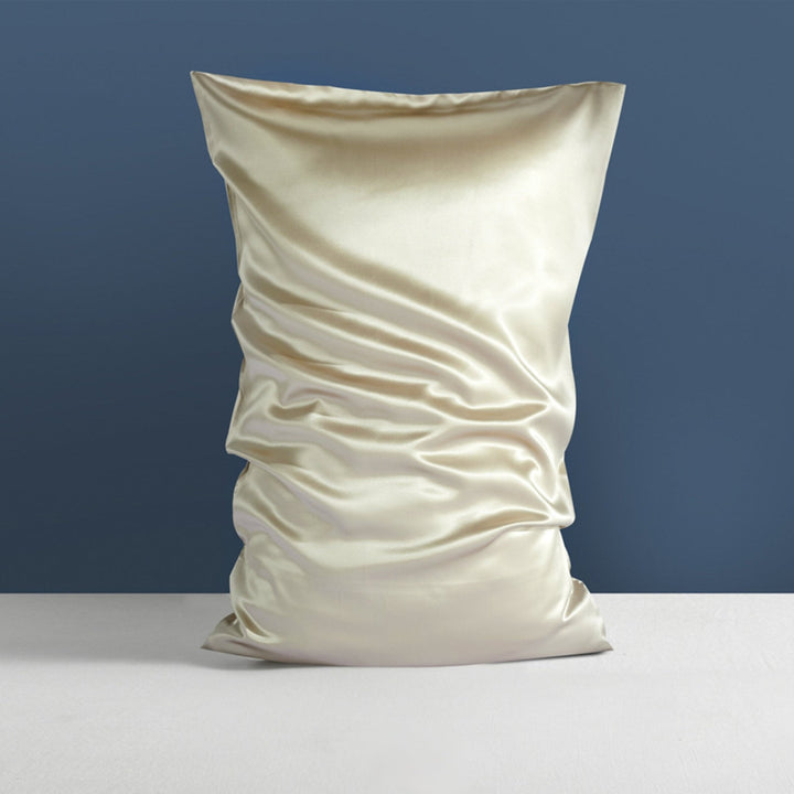 A wrinkled, cream-colored 22 Momme High Gloss Silk Pillowcase Envelope by SusanSilk stands upright against a grayish-blue wall on a white surface.