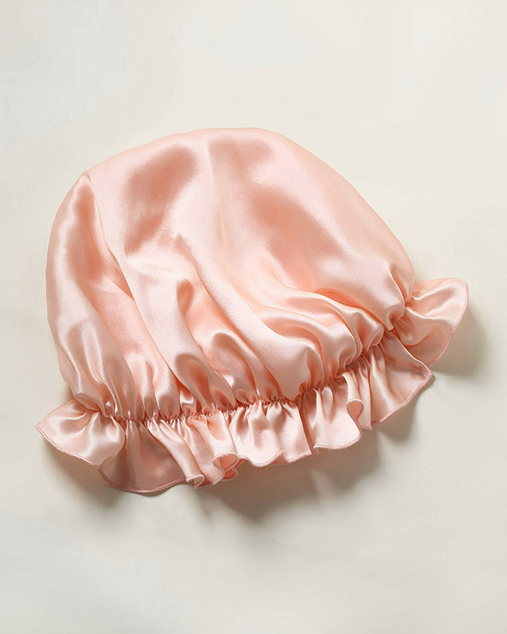 A Flounced Silk Bonnet For Sleeping by SusanSilk, featuring pink satin and ruffled edges, commonly used for hair protection during sleep, is laid flat on a light-colored surface.