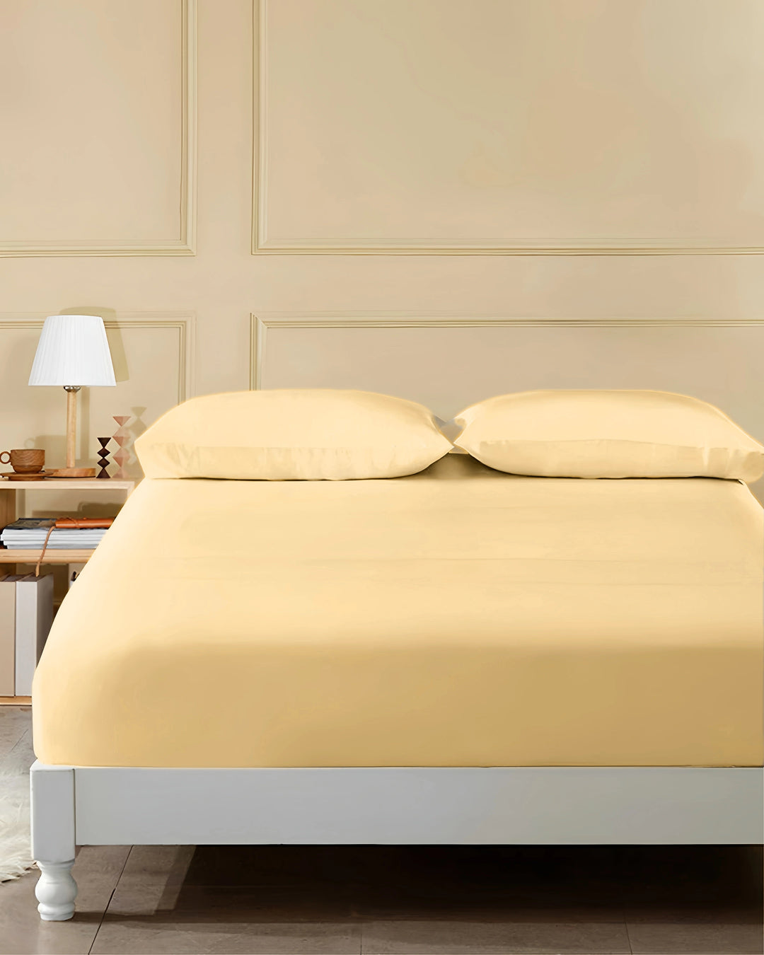Beige bedspread on a bed in a bedroom setting with a lamp and books on a shelf.