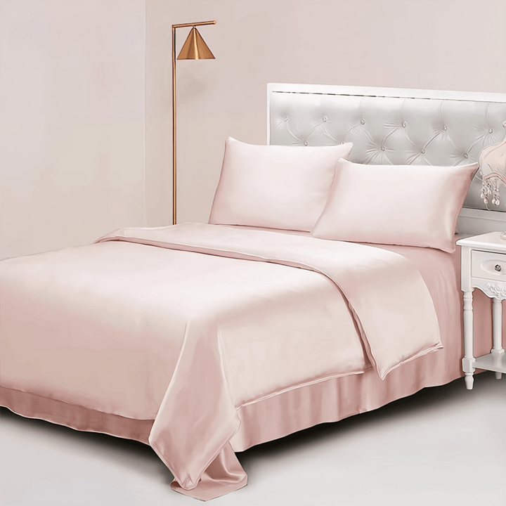 A neatly made bed adorned with the 25 Momme Luxury Silk Bedding Set - 4Pcs from SusanSilk in light pink, complemented by a tufted white headboard and a gold floor lamp beside it. A small white bedside table with a floral decoration is also visible, exuding elegance and charm with its SusanSilk touch.