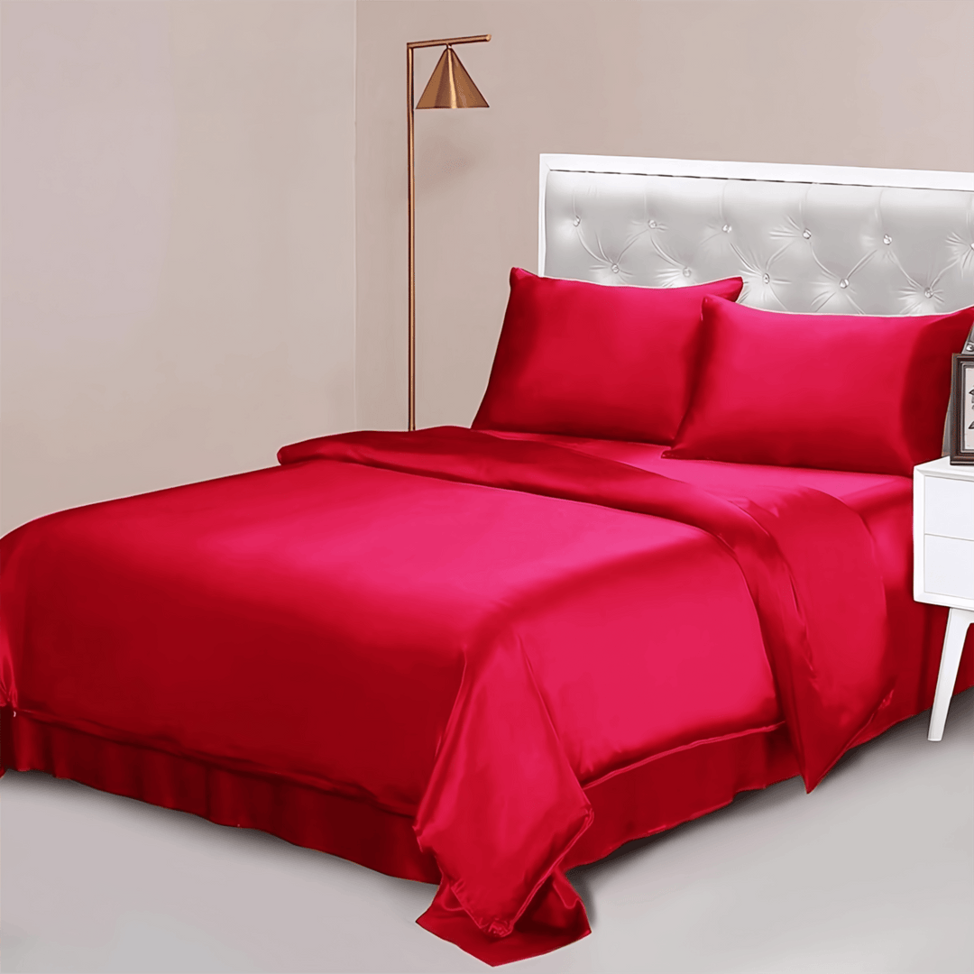 A neatly made bed with the 25 Momme Luxury Silk Bedding Set - 4Pcs by SusanSilk, featuring vibrant red silk comforter, sheets, and pillowcases. The bed’s white tufted headboard is set against a beige wall, complemented by a standing lamp and nightstand nearby.