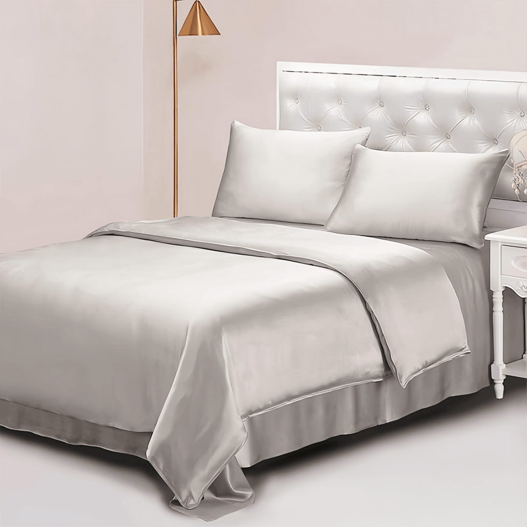 A neatly made bed featuring the 25 Momme Luxury Silk Bedding Set - 4Pcs by SusanSilk in a sophisticated gray, complemented by a stylish white tufted headboard and matching silk pillows. Beside the bed sits a white nightstand adorned with a decorative item, and in the corner stands an elegant floor lamp with a gold shade.