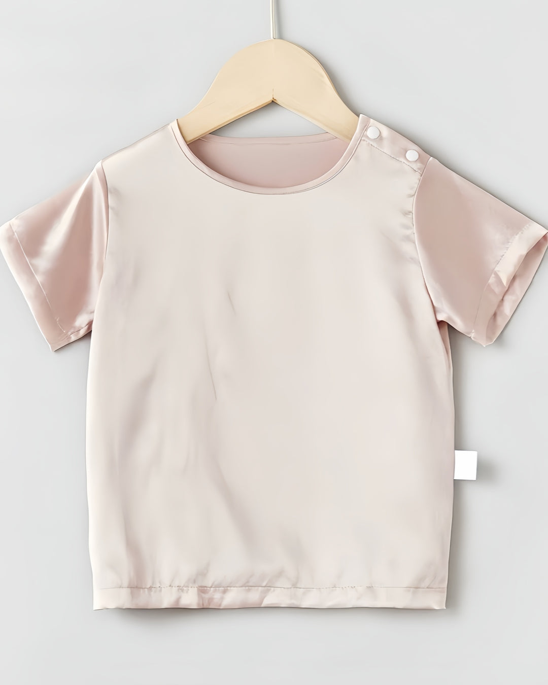 A delicate pink short-sleeved silk top from SusanSilk, crafted in luxurious 19 Momme Mulberry Silk, gently hangs on a wooden hanger against a plain background. Its fabric and two shoulder buttons evoke the softness of a baby's second skin.