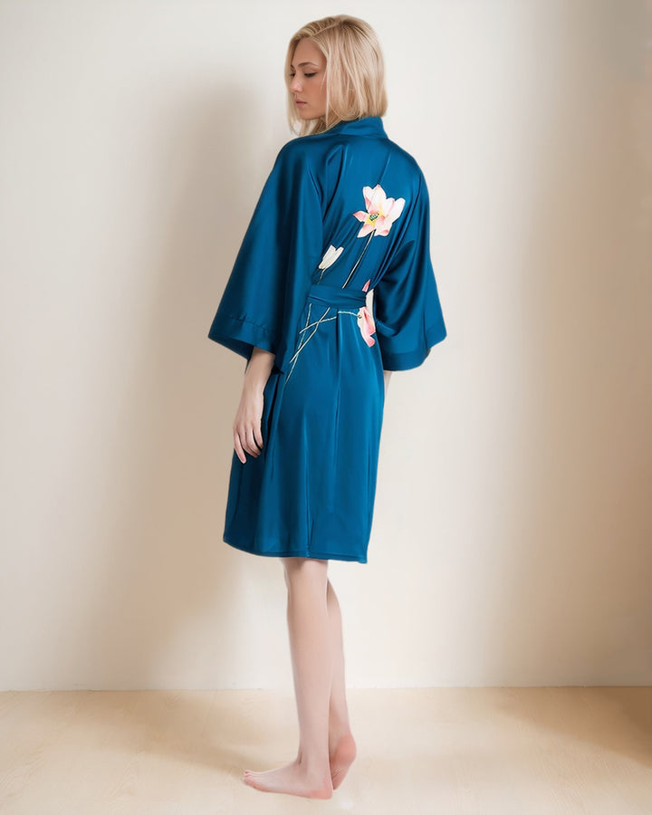 A person with long, blonde hair stands barefoot wearing the Lotus Style Short Silk Kimono by SusanSilk. They are facing away, slightly turning their head to the left, showcasing the blue robe with floral designs on the back.