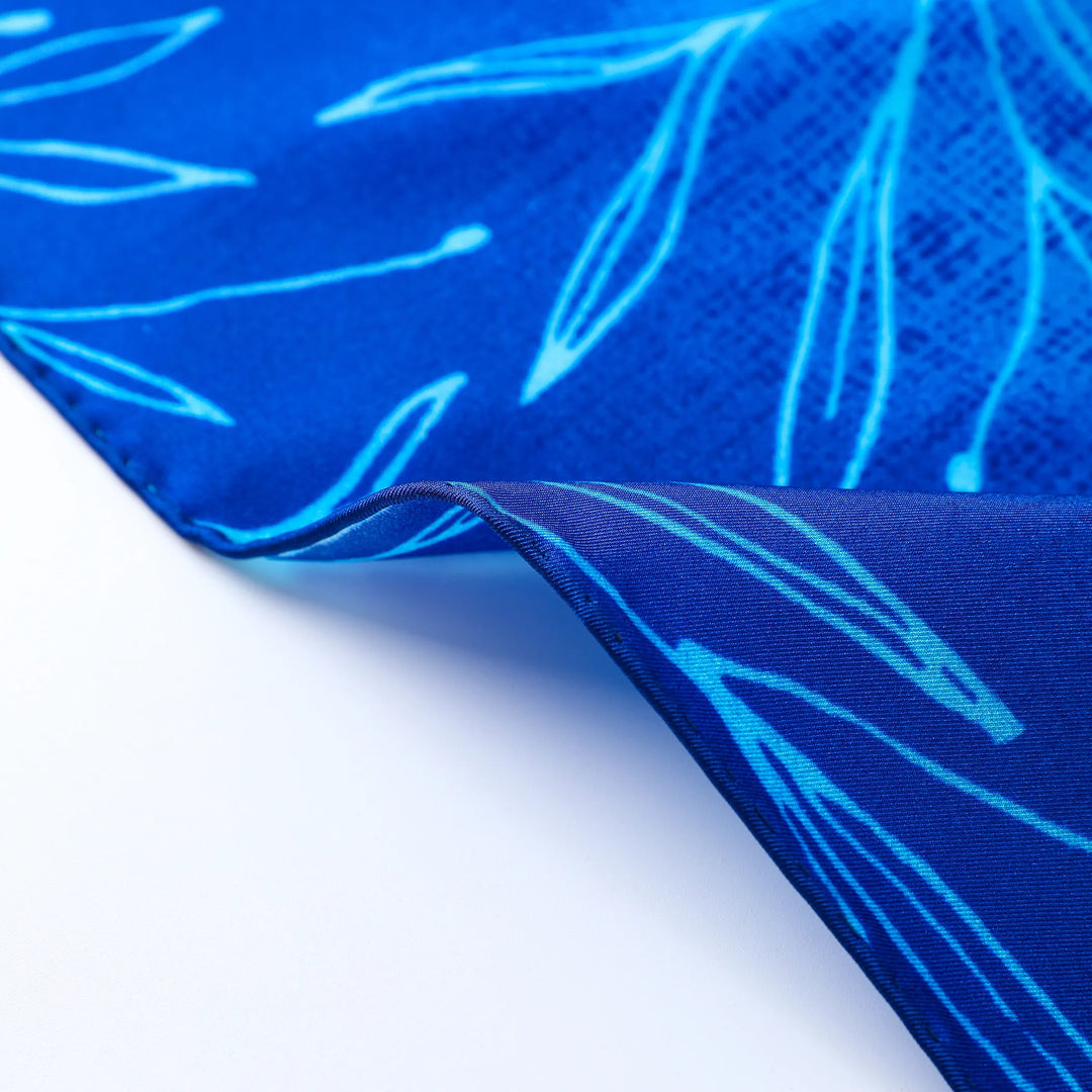 Close-up of a section of the Blue Sky Square Silk Scarf by SusanSilk, featuring light blue leaf patterns. The image shows a corner slightly folded over, revealing the underside with the same intricate design.