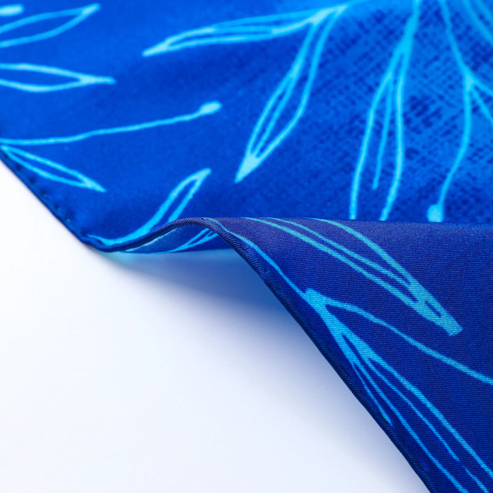 Close-up of a section of the Blue Sky Square Silk Scarf by SusanSilk, featuring light blue leaf patterns. The image shows a corner slightly folded over, revealing the underside with the same intricate design.