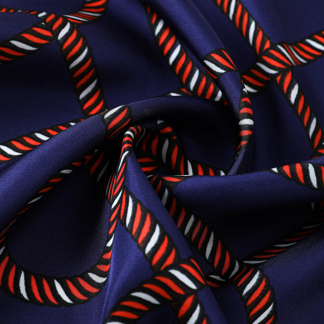 Close-up of the SusanSilk Witch Hazel Orange Square Silk Scarf, showcasing a navy blue fabric with a twisted rope pattern in red, white, and black, arranged in a geometric design. The silk fabric appears slightly folded.