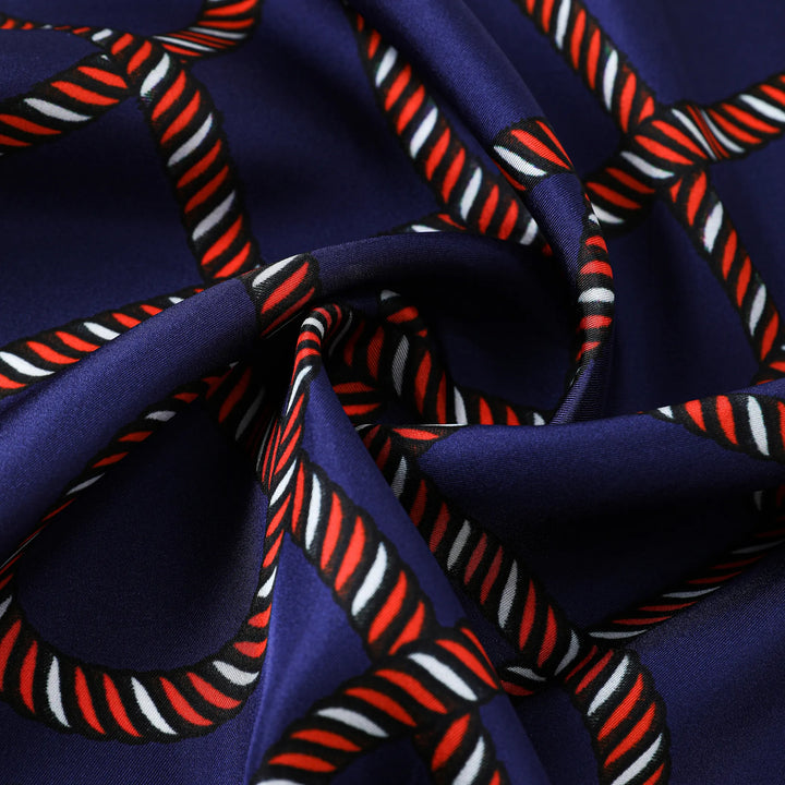 Close-up of the SusanSilk Witch Hazel Orange Square Silk Scarf, showcasing a navy blue fabric with a twisted rope pattern in red, white, and black, arranged in a geometric design. The silk fabric appears slightly folded.