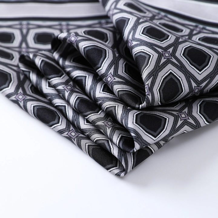 The Sakuma Gray Square Silk Scarf by SusanSilk showcases folded silk fabric adorned with a geometric pattern in shades of black, gray, and white, featuring hexagonal shapes and parallel lines.