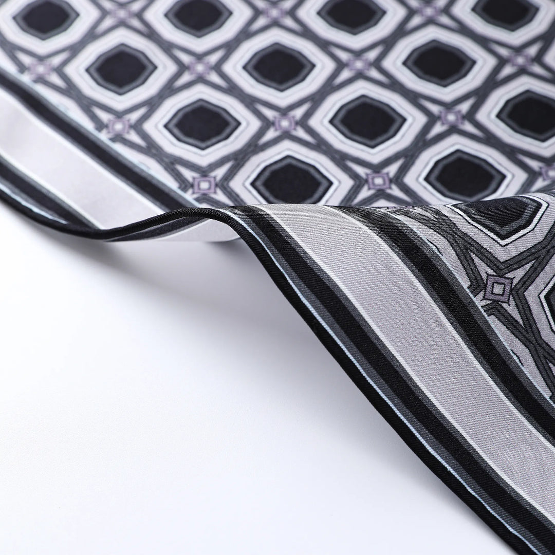 Close-up of the Sakuma Gray Square Silk Scarf by SusanSilk, showcasing a geometric diamond pattern in black, white, and light purple. The luxurious fabric is further accentuated by a contrasting striped border.