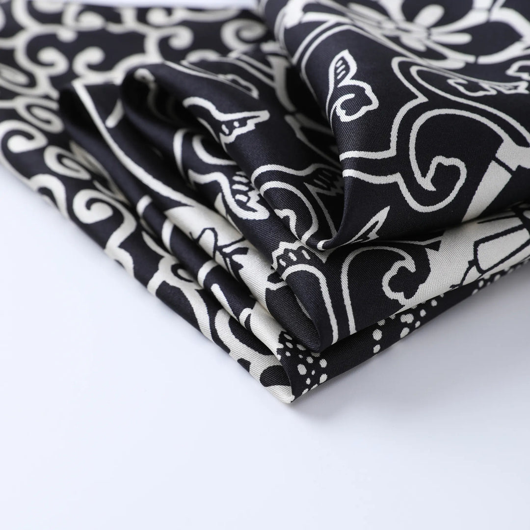 A close-up view of the Seeker Black Square Silk Scarf by SusanSilk, showcasing its intricate, swirling patterns in black and white, folded neatly on a white surface.