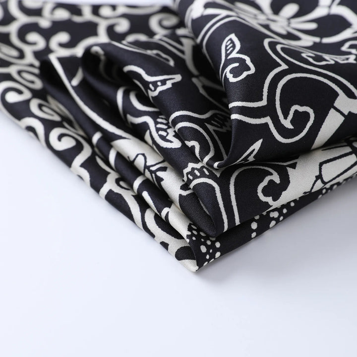 A close-up view of the Seeker Black Square Silk Scarf by SusanSilk, showcasing its intricate, swirling patterns in black and white, folded neatly on a white surface.