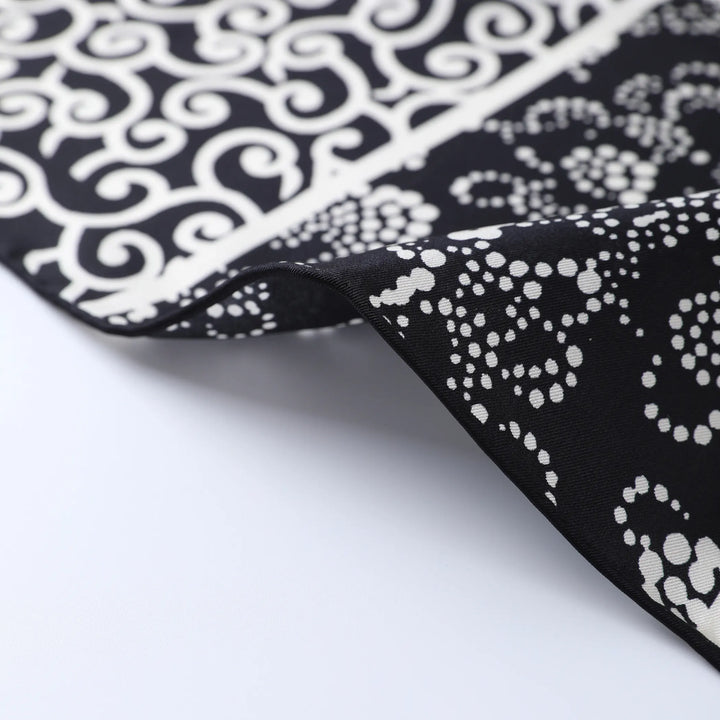 Close-up of the luxurious Seeker Black Square Silk Scarf by SusanSilk, showcasing its elegant black and white patterns. The upper portion displays a white swirling design on a black background, while the lower section features intricate white dotted motifs on the same dark backdrop.