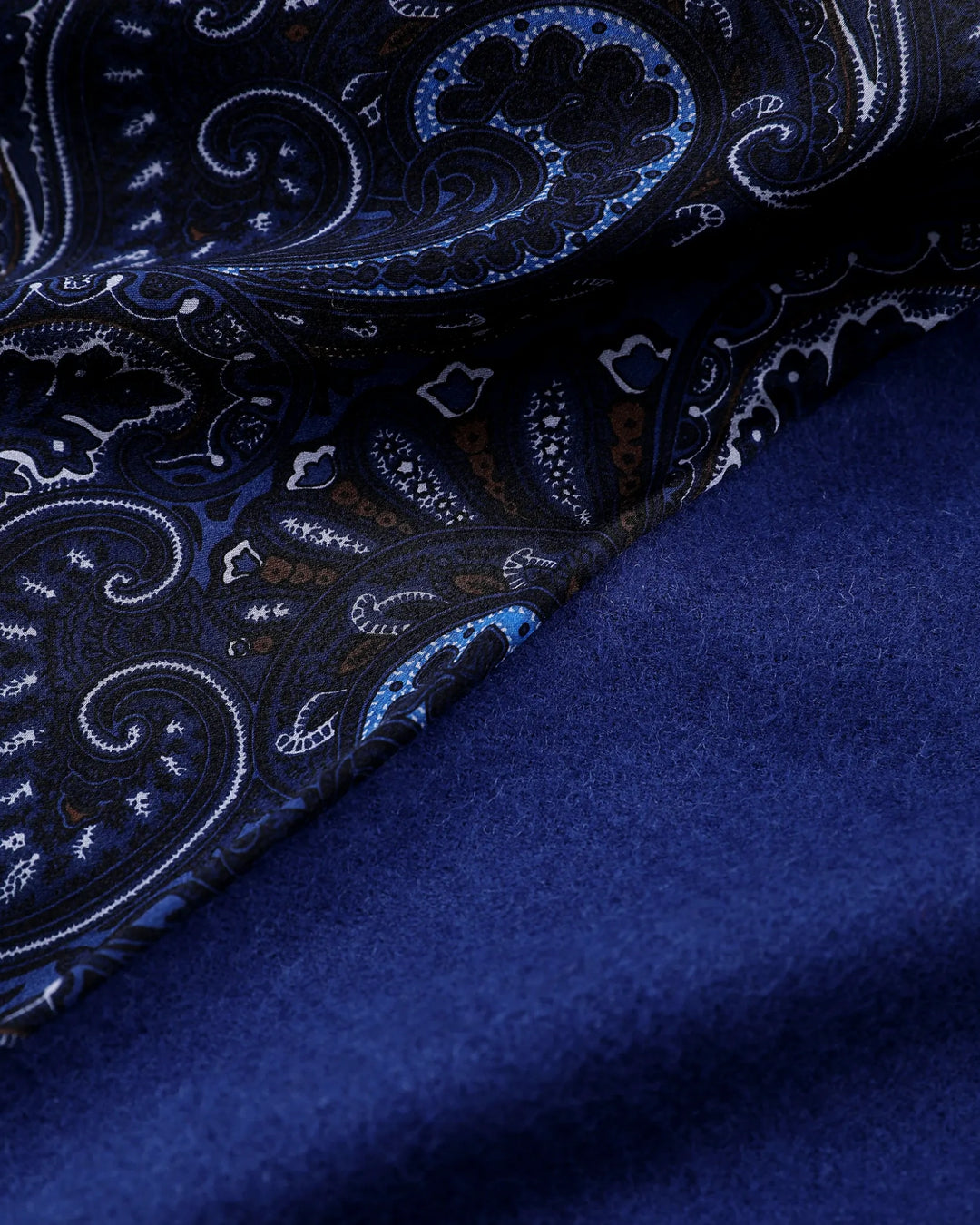 A close-up of a folded fabric item, likely a scarf or shawl, showing a dark navy blue base with an intricate paisley pattern in lighter blue, white, and subtle brown accents, layered over a solid deep blue velvet-like section.#color_navy-blue