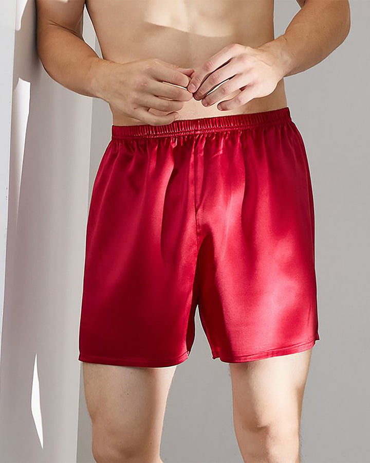 A person stands indoors wearing SusanSilk's 19 Momme Comfortable Silk Boxer in red, their hands positioned near the waist, and no visible shirt. The sheen of the fabric catches the light beautifully.