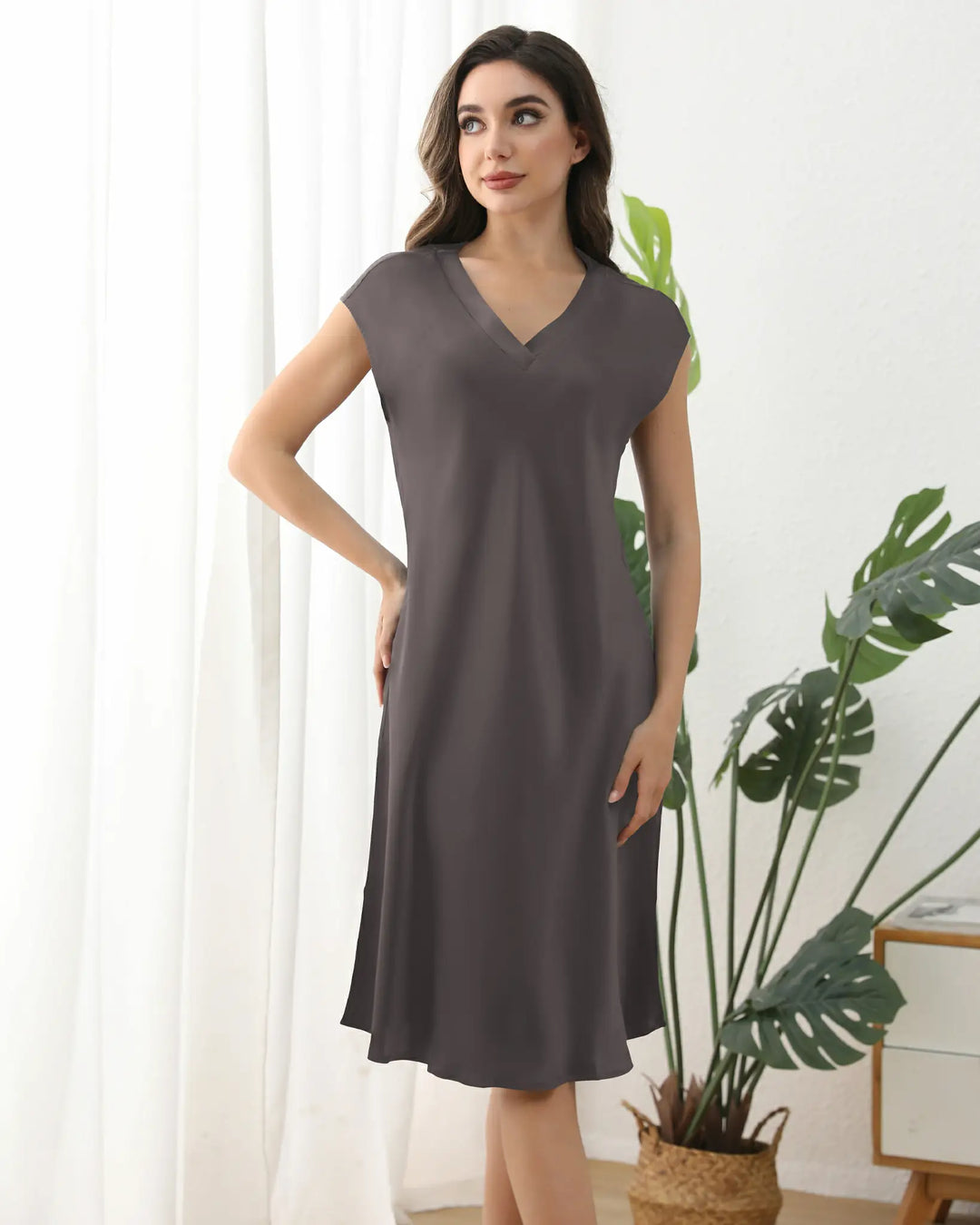 A woman stands against a plain background, wearing the SusanSilk 22 Momme Comfortable Silk Nightdress. This simple, knee-length, short-sleeved gray dress features a V-neck and is made of soft silk that elegantly drapes over her figure. She has long, straight hair and is looking down with a slight smile.#color_dark-grey