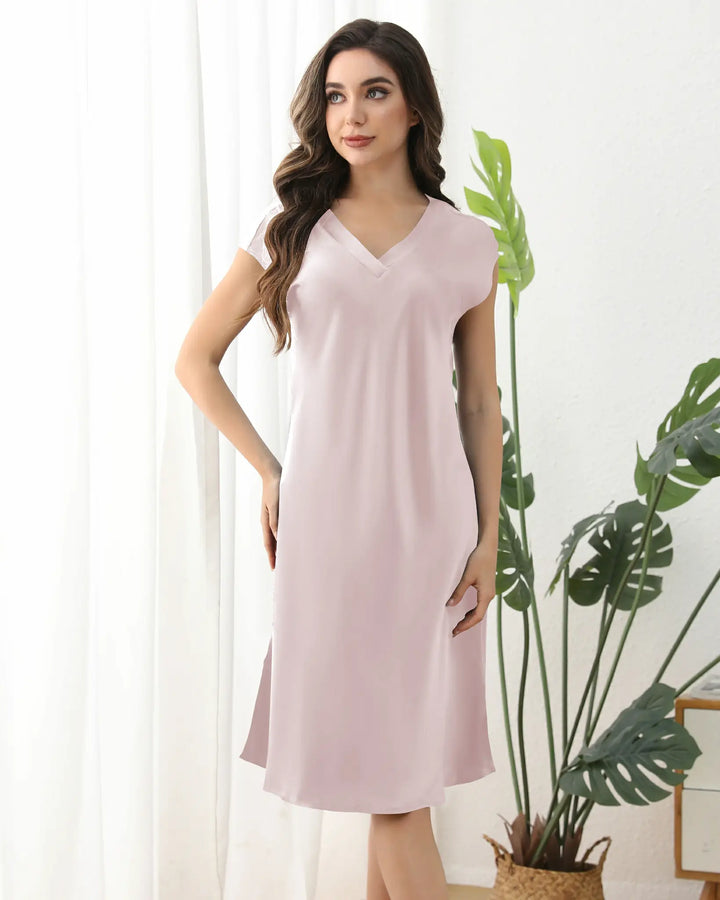 A woman wearing a SusanSilk 22 Momme Comfortable Silk Nightdress in light pink stands against a plain grey background, smiling slightly with her right hand touching the side of her face.#color_light-pink