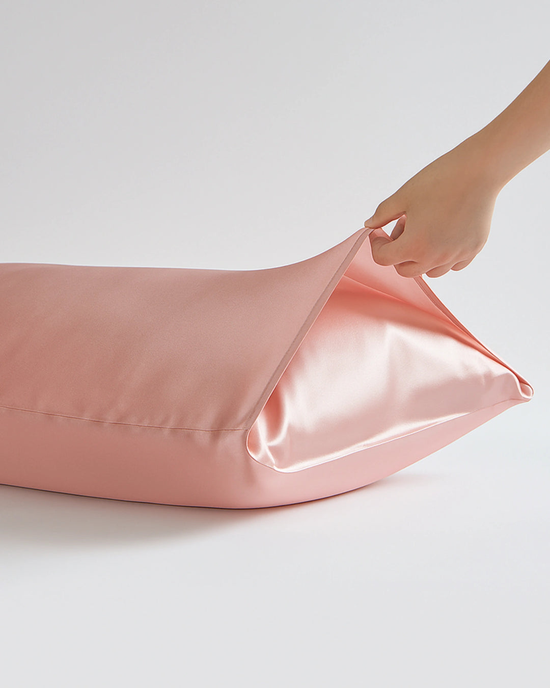 Pink satin pillowcase being adjusted by a hand on a light gray background