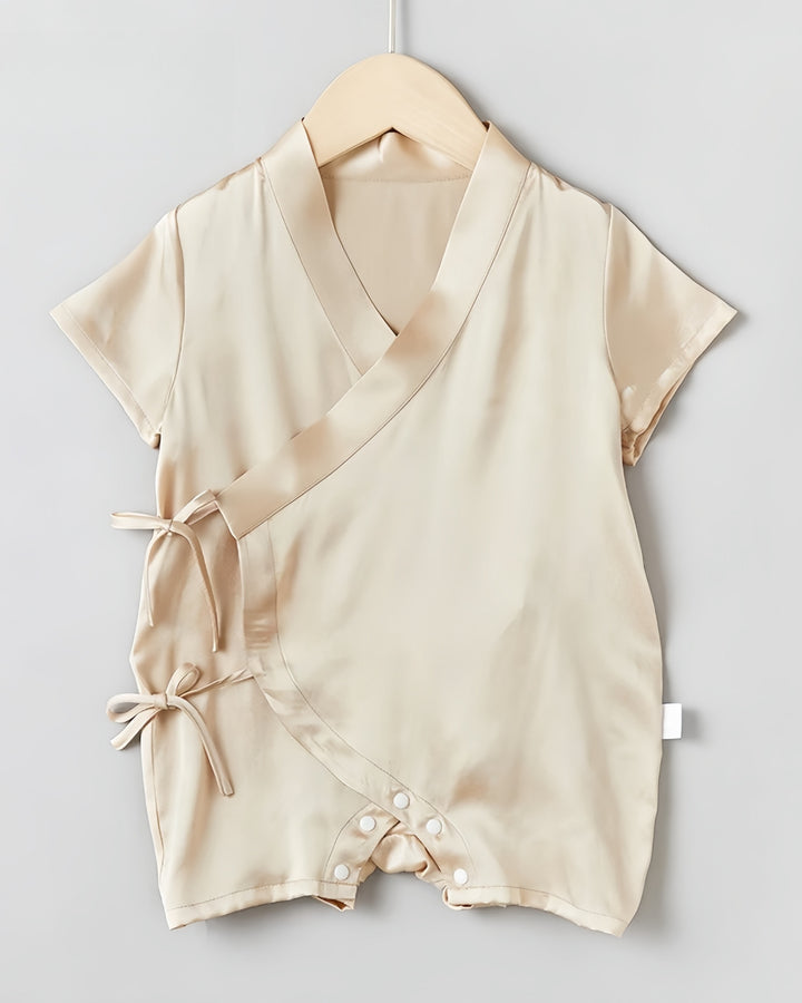The 19 Momme Silk Baby Button Lace-Up Onesie from SusanSilk, a beige short-sleeve romper featuring a wrap-style front with two side ties and snap closures at the crotch, is made from silky smooth fabric and hangs on a wooden hanger against a light gray background.