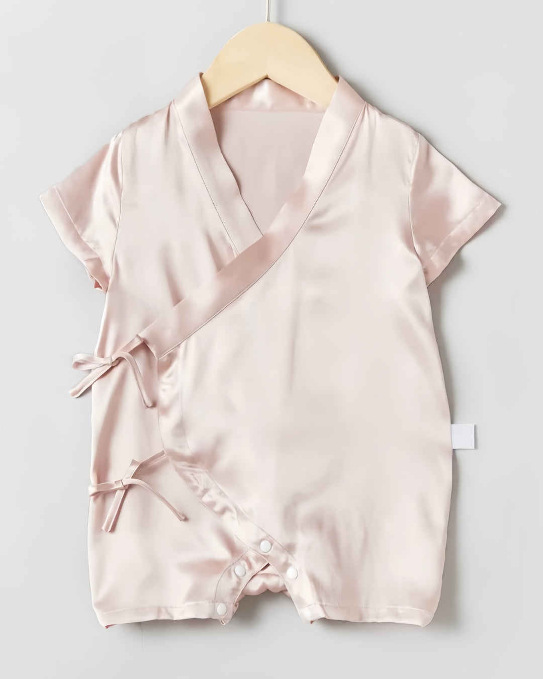 The 19 Momme Silk Baby Button Lace-Up Onesie by SusanSilk is a light pink satin baby romper featuring short sleeves, a crossover front tied with bows, and snap closures at the bottom. It is showcased on a wooden hanger against a neutral background.