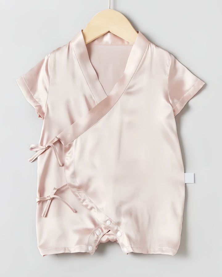 The 19 Momme Silk Baby Button Lace-Up Onesie by SusanSilk is a light pink satin baby romper featuring short sleeves, a crossover front tied with bows, and snap closures at the bottom. It is showcased on a wooden hanger against a neutral background.