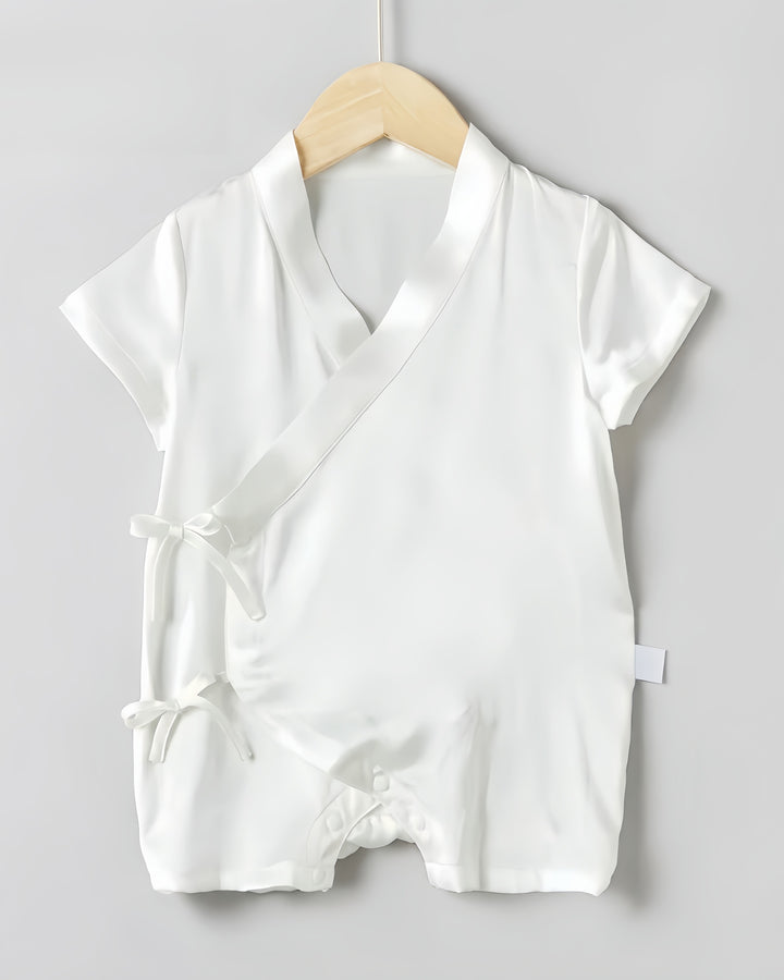 A 19 Momme Silk Baby Button Lace-Up Onesie by SusanSilk, featuring a white, short-sleeved design with adjustable side ties and snap buttons at the bottom, hangs on a wooden hanger against a plain gray background.