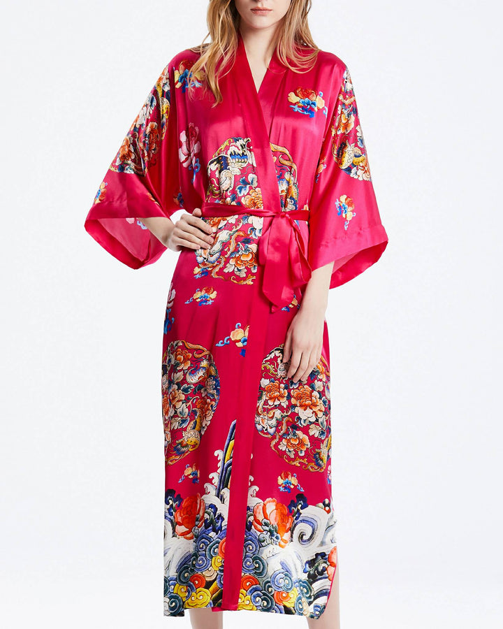 A woman stands wearing the Wheel of Fortune Long Silk Kimono, a vibrant red silk robe adorned with intricate, colorful patterns and a waist sash from SusanSilk.#color_rose-red