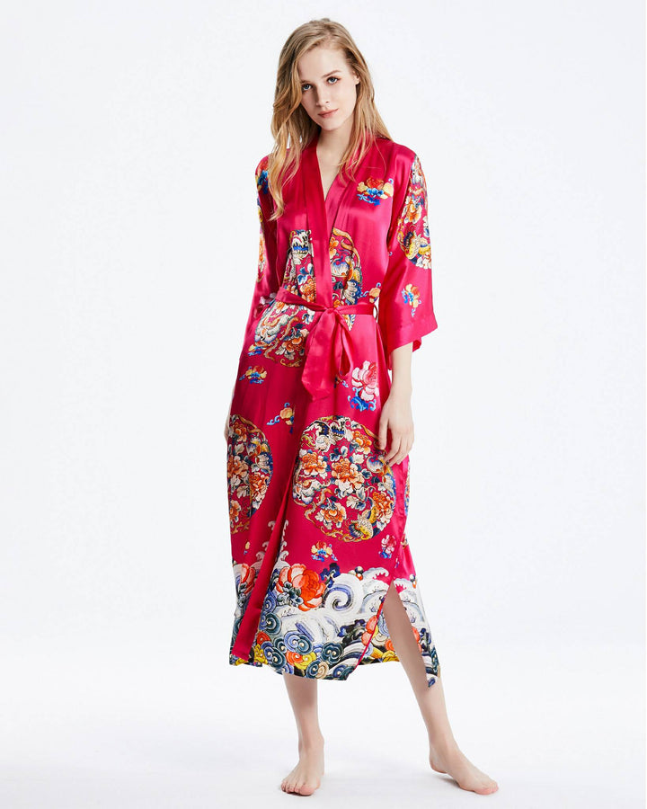 A woman stands barefoot wearing the Wheel of Fortune Long Silk Kimono, a vibrant, floor-length red robe with intricate, colorful patterns from the prestigious SusanSilk brand. The luxurious kimono features a belt tied at the waist and wide sleeves.#color_rose-red