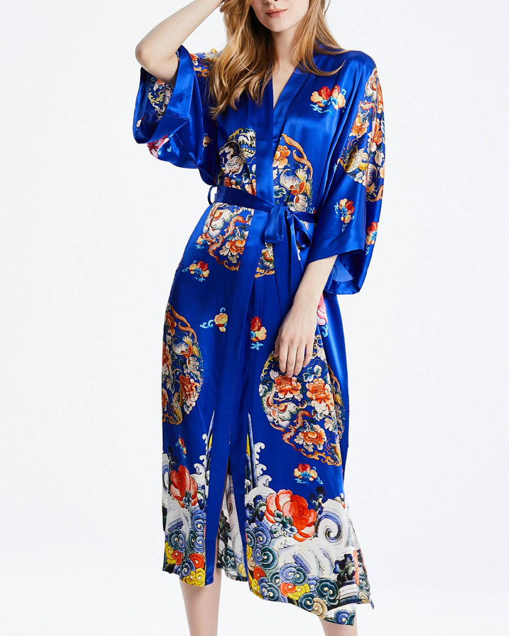 A woman wearing a Wheel of Fortune Long Silk Kimono from SusanSilk, featuring intricate, colorful floral and wave patterns, stands against a plain white background.#color_royal-blue