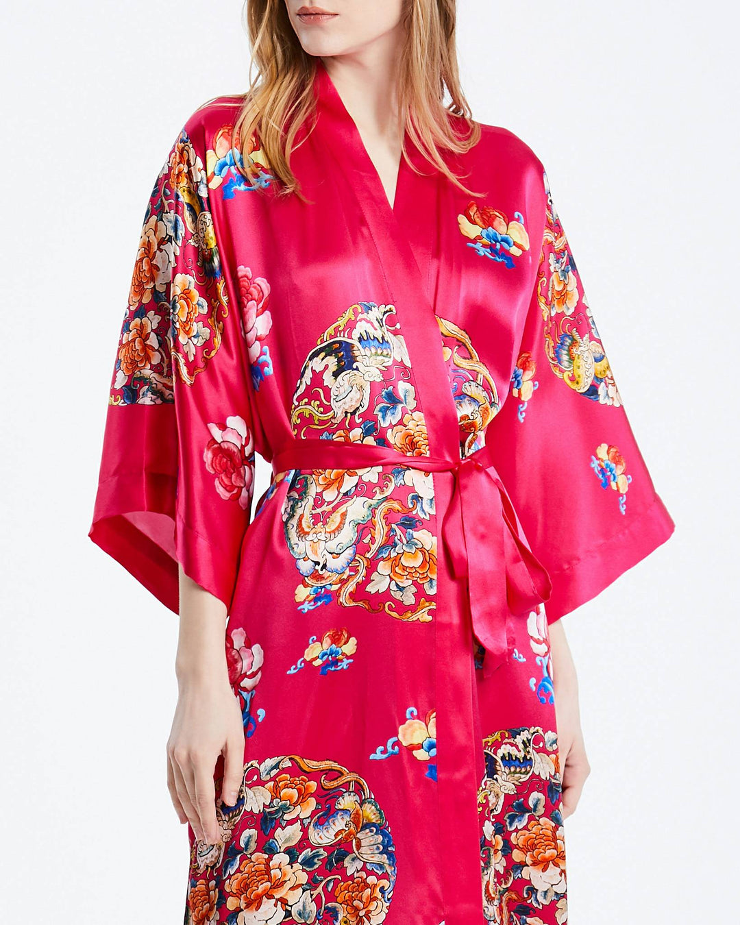 A person wearing the Wheel of Fortune Long Silk Kimono by SusanSilk, featuring vibrant red fabric adorned with intricate multicolored floral and dragon patterns, stands against a plain white background.#color_rose-red