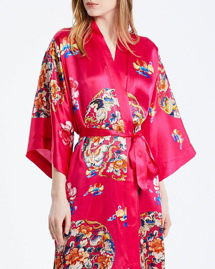 A person wearing the Wheel of Fortune Long Silk Kimono by SusanSilk, featuring vibrant red fabric adorned with intricate multicolored floral and dragon patterns, stands against a plain white background.#color_rose-red