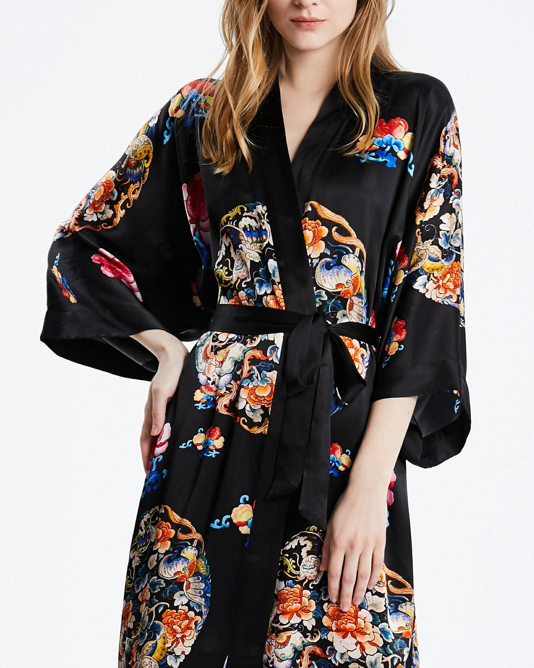 A person wearing the Wheel of Fortune Long Silk Kimono by SusanSilk, adorned with colorful floral patterns, stands with one hand on their hip. The black silk kimono features wide sleeves and a delicate waist tie.#color_black