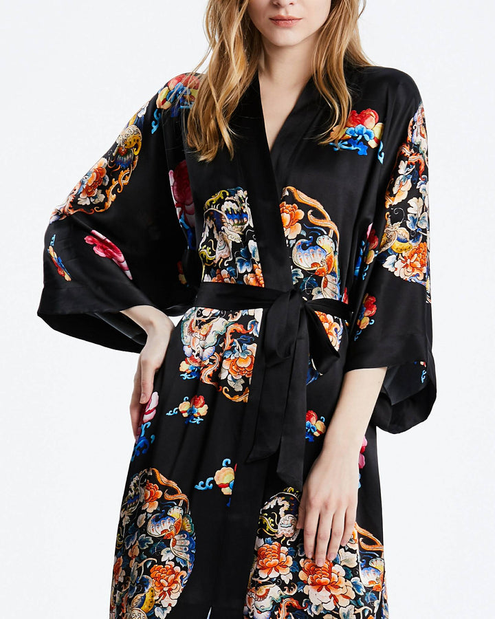 A person wearing the Wheel of Fortune Long Silk Kimono by SusanSilk, adorned with colorful floral patterns, stands with one hand on their hip. The black silk kimono features wide sleeves and a delicate waist tie.#color_black