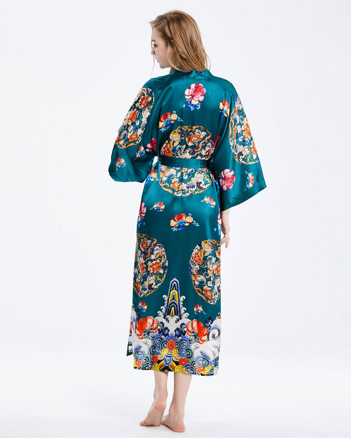 A woman stands barefoot, facing away, wearing a turquoise Wheel of Fortune Long Silk Kimono by SusanSilk, adorned with colorful floral and traditional patterns.#color_sea-green