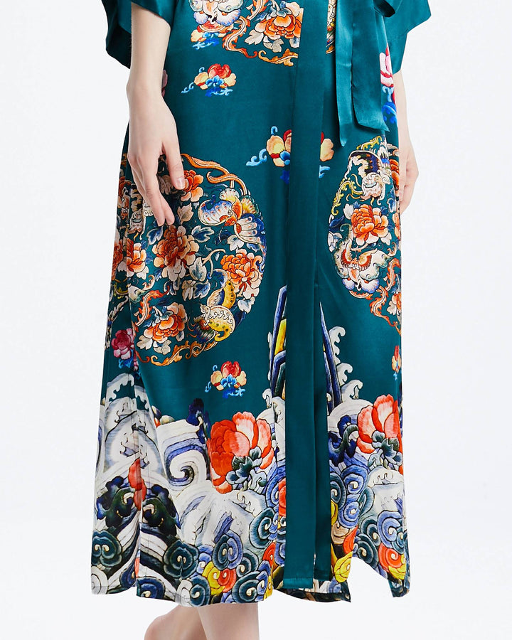 Torso and lower body of a person wearing a vibrant, patterned Wheel of Fortune Long Silk Kimono by SusanSilk featuring floral and abstract designs in various colors on a teal background. The person stands against a plain, light backdrop.#color_sea-green