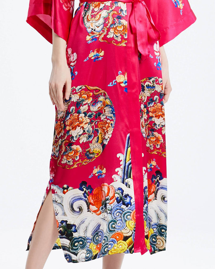 A person wearing the Wheel of Fortune Long Silk Kimono by SusanSilk, showcasing colorful and intricate floral and wave patterns on luxurious red susansilk. The kimono features a side slit and a fabric belt.#color_rose-red